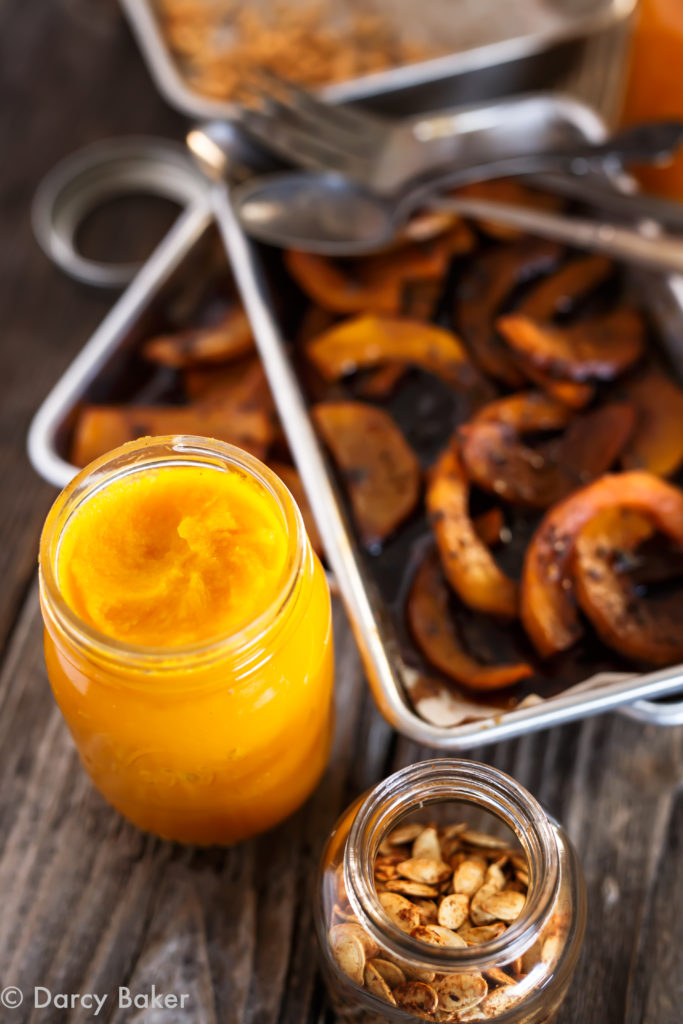 jar of roasted squash puree in front, slightly top down view with soft focus and trays slightly off behind stacked diagonally with pieces of roasted squash.  A small jar or roasted seeds in the front, slightly cut off in image on a dark wooden surface