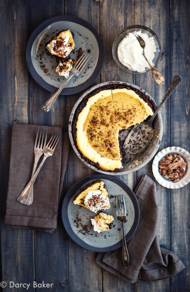 top down view of squash cheese cake with whip cream, top down view on dark grey background with a tan linen napkin a silver serving spoon, two plates with slices, brown linen napkins, a bowel of pecans and a bowl of fresh whipped cream